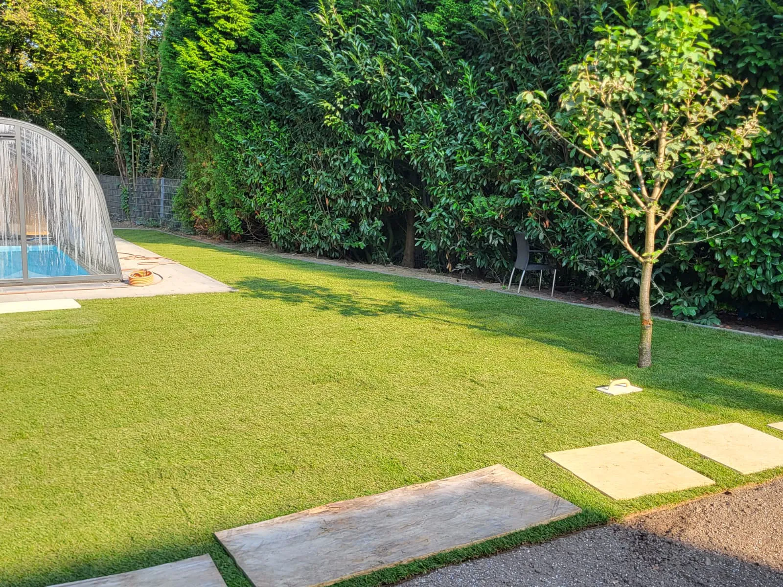 Rasenfläche mit frischem grünem Rasen, Natursteinplatten als Trittsteine und teilabgedecktem Pool im Garten