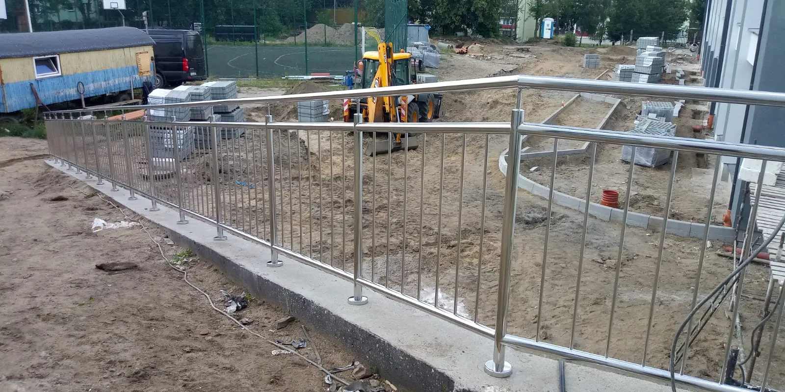 Metallgeländer aus Edelstahl auf massivem Betonsockel vor einer Baustelle mit Erdarbeiten und Bagger