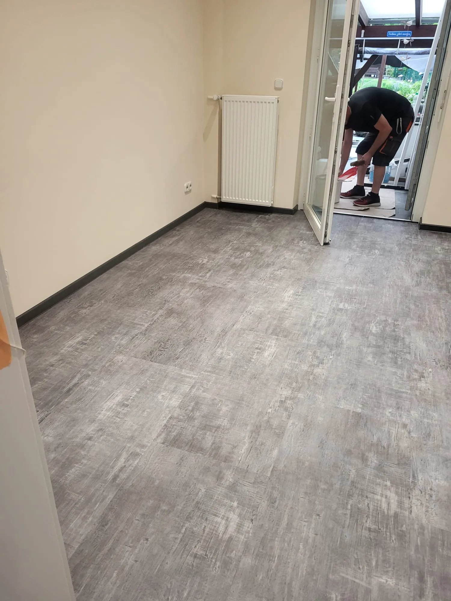 Vinylboden verlegen: grauer Boden in Innenraum, Handwerker fegt Boden am offenen Türbereich